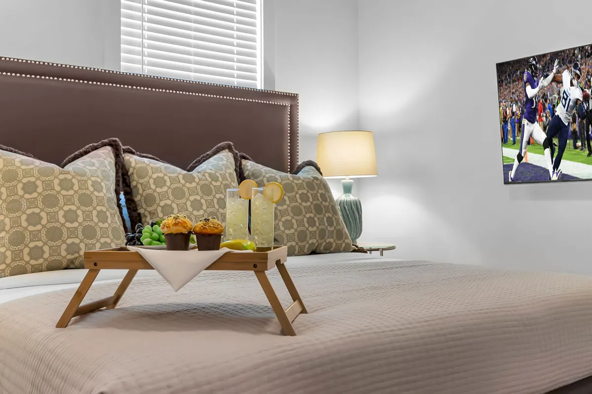 A spacious bedroom with a large bed adorned with decorative pillows, a breakfast tray with drinks and pastries, a warm lamp, and a TV showing a sports event. The mood is cozy yet lively, suggesting comfort and entertainment in one private space.