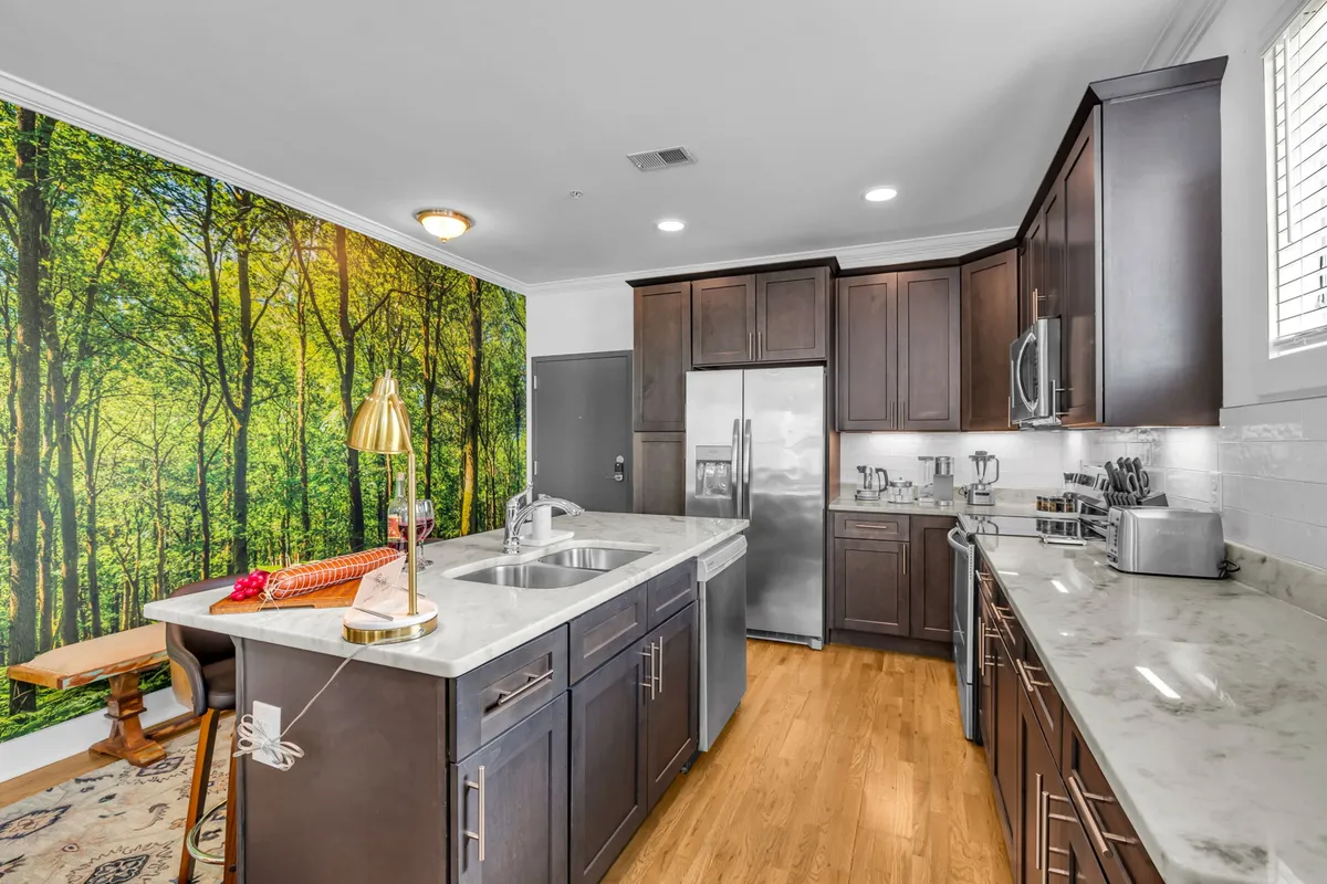 This image captures the heart of the open-concept living, dining, and kitchen area. It features a kitchen island with a marble countertop, stainless-steel appliances, and the standout forest mural that defines the space. The inviting arrangement hints at both relaxation and social gatherings.