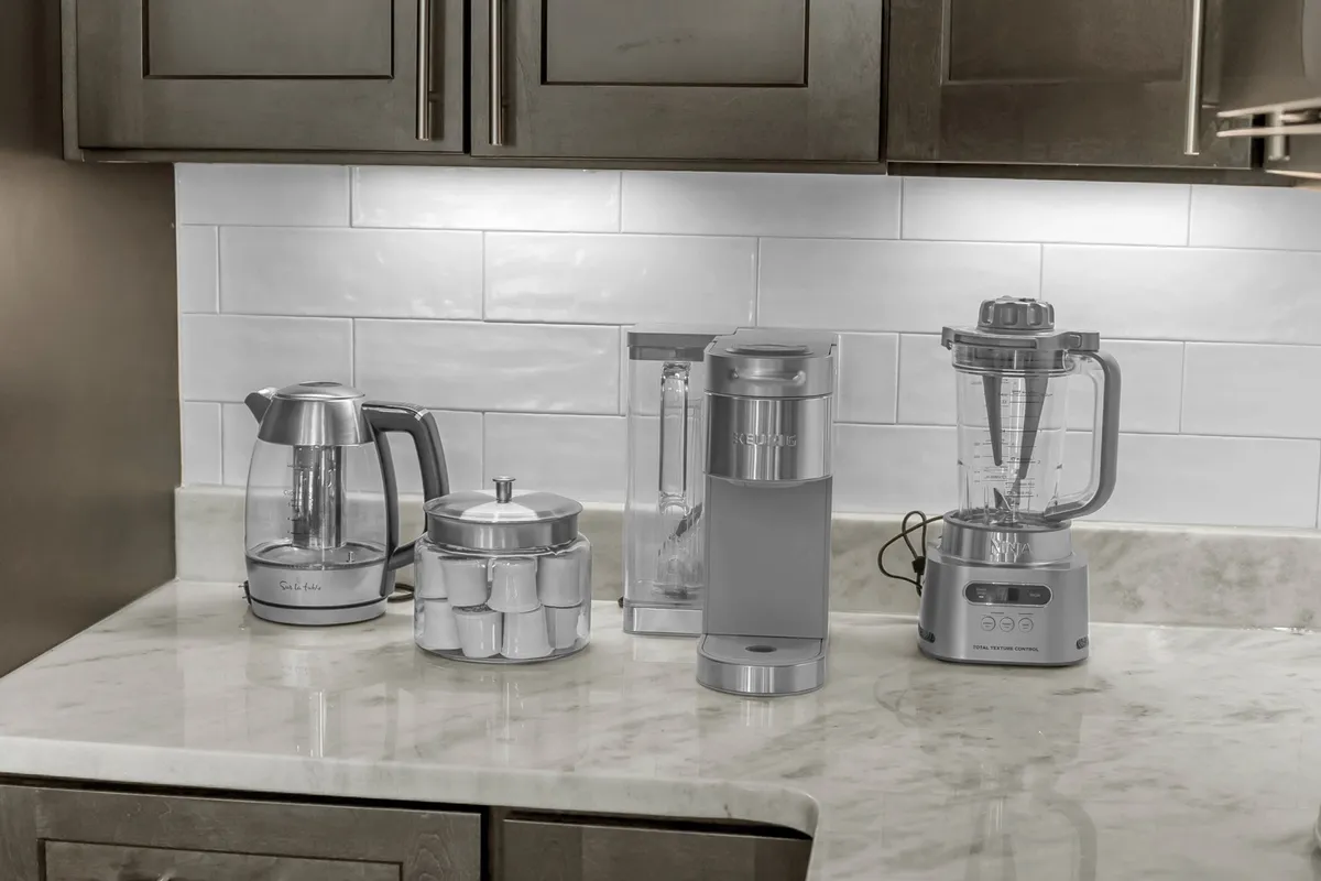 A tidy beverage station with an electric kettle, coffee maker, water dispenser, blender, and a canister of coffee pods, all arranged on a countertop. The setup is functional, convenient, and ready to use on arrival.