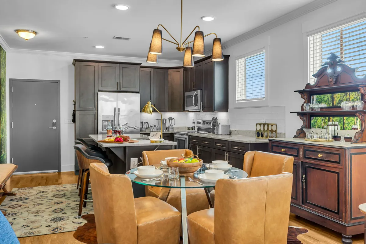 A modern open-plan kitchen and dining area featuring sleek appliances, a round table with comfortable seating, contrasting cabinetry, and a decorative serving cart. The space is bright, inviting, and designed for both social gatherings and everyday convenience.