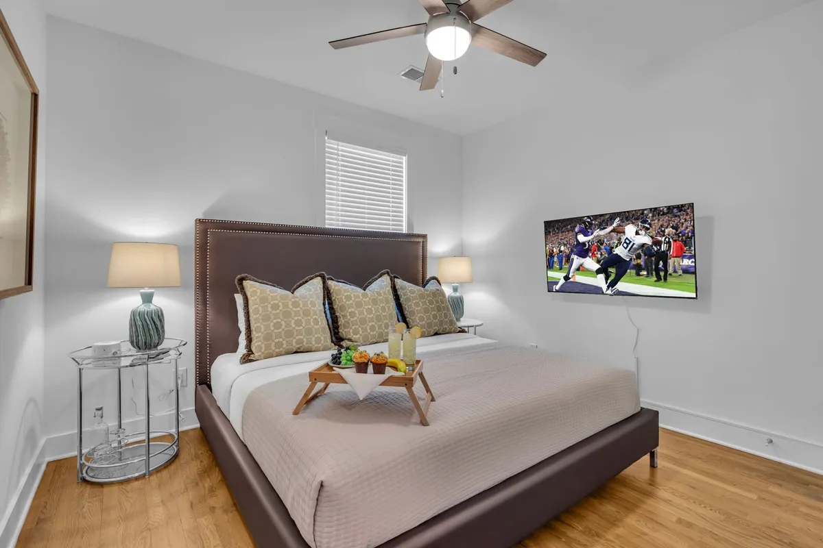 A cozy king bedroom featuring a large bed adorned with decorative pillows, nightstands with soft-glowing lamps on each side, a TV mounted on the wall, and a tray of snacks inviting relaxation on the bed. The calming décor and gentle lighting create an inviting and restful environment.