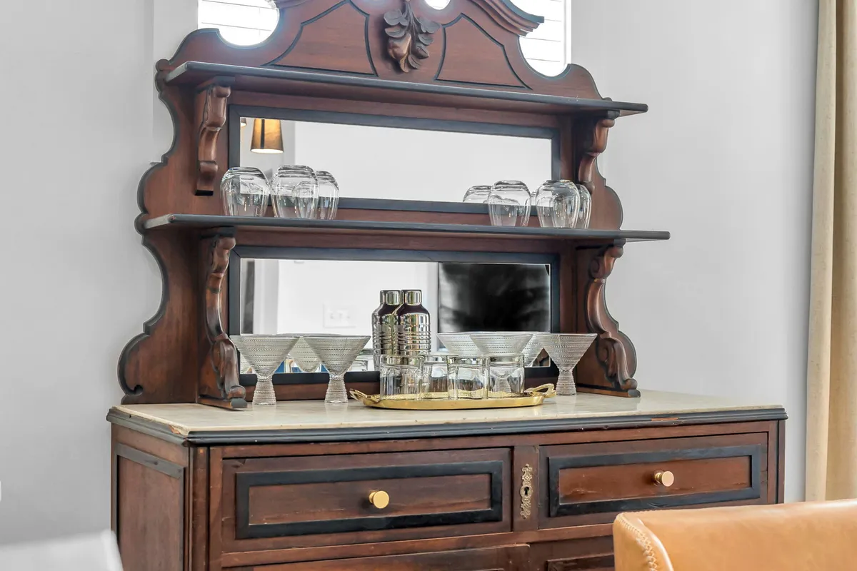A sophisticated hutch area is displayed, with ornate wooden cabinetry, glassware, a mirror, and a golden tray ready for mixing drinks. The elegant setup hints at a space dedicated to hosting and enjoyment.