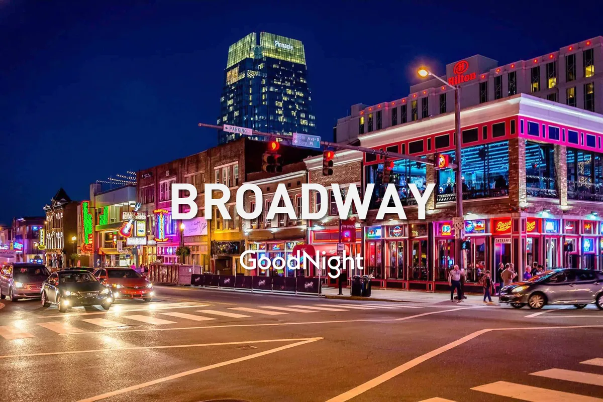 A bustling downtown Nashville street at night, lined with colorful neon signs, busy sidewalks full of people, and a tall building in the background. The energetic scene captures the excitement of live music venues, restaurants, and the city's famous nightlife atmosphere.