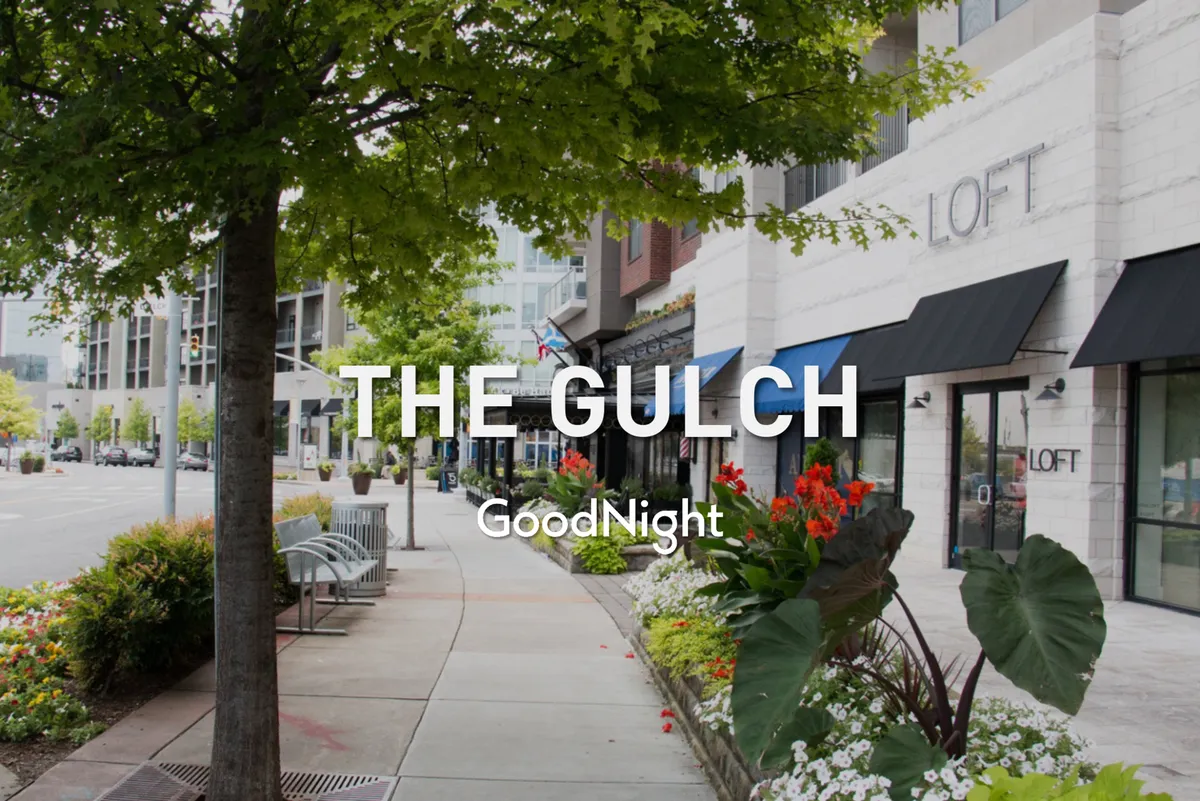 A vibrant, tree-lined sidewalk flanked by flower beds and storefronts. Benches offer spots to relax, and the scene is lively, inviting, and full of local character, suggesting a walkable area with outdoor spaces and pet-friendly cafés.